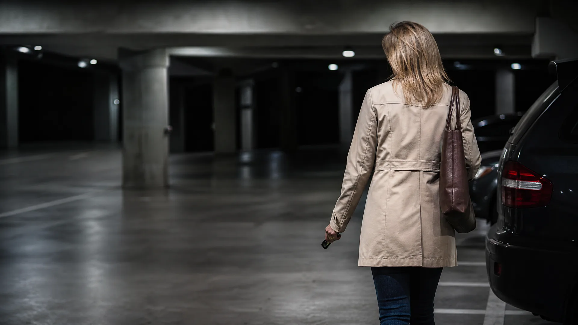 A woman in a beige coat with a brown shoulder bag walks away from the camera through a dimly lit concrete parking garage, holding keys, with a dark parked car on the right.